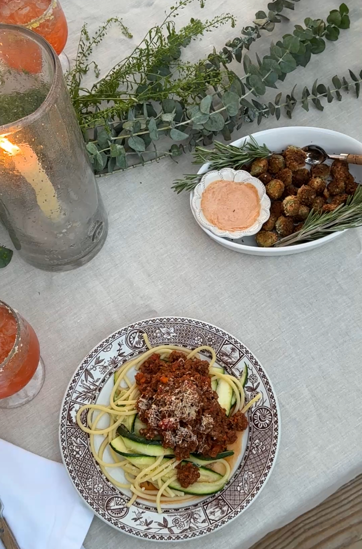 A detailed shot of a pasta dish with ragu and zucchini ribbons on a vintage patterned plate, set alongside fried stuffed olives with rosemary sprigs and a tall flickering candle.