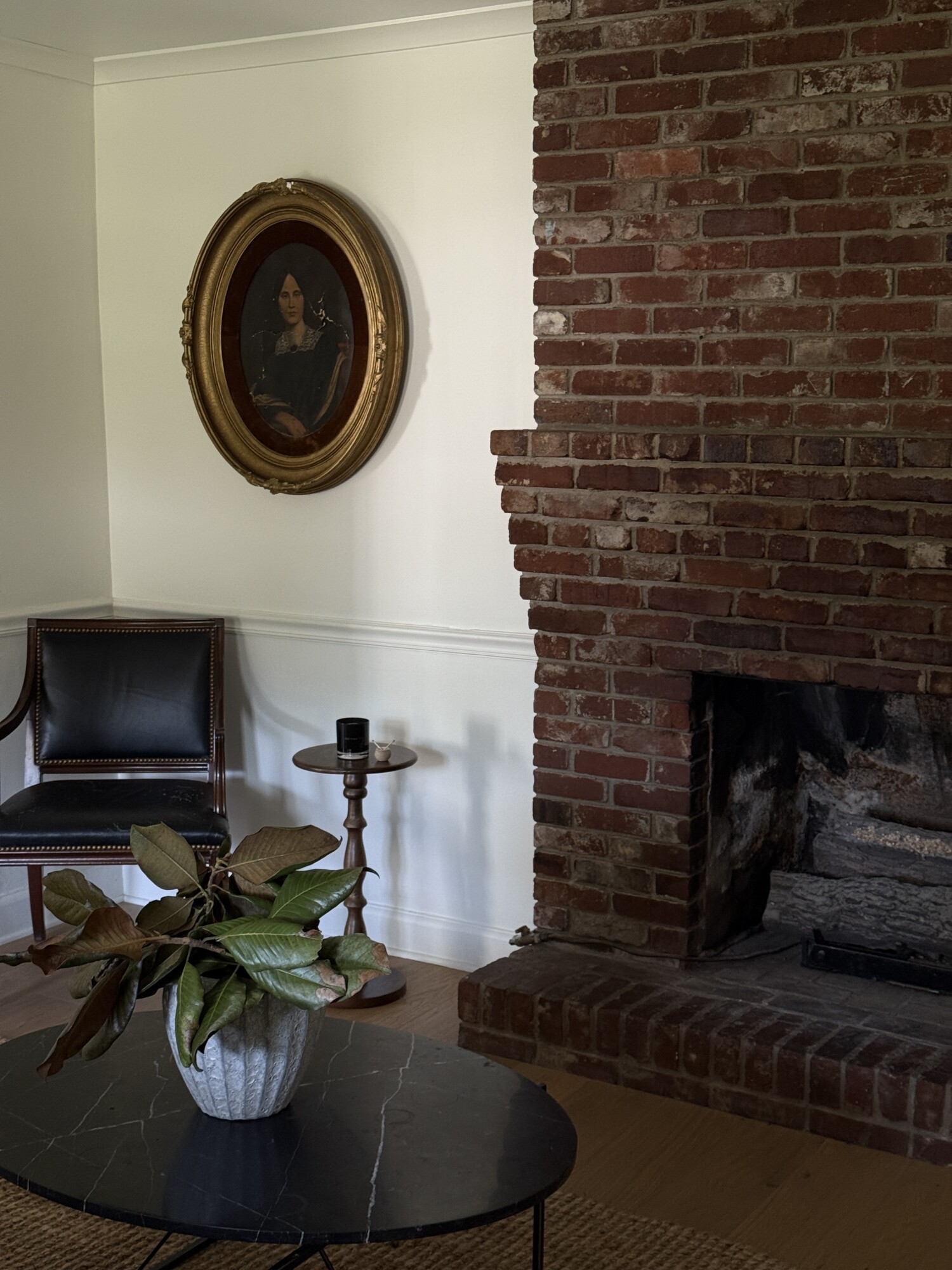Cozy living room corner with exposed red-brick fireplace, black leather chair with nailhead trim, marble coffee table with a magnolia plant, and an antique gilt oval portrait.