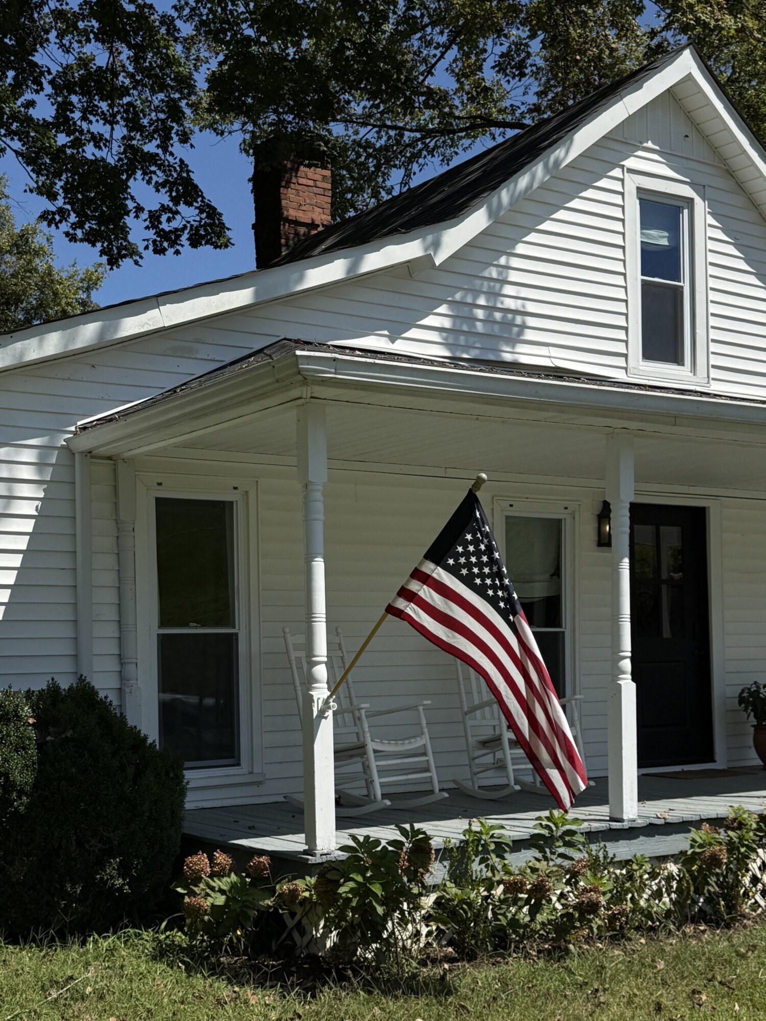 White clapboard farmhouse with front porch, two white rocking chairs, and an American flag, bordered by hydrangeas and mature trees on a sunny day.