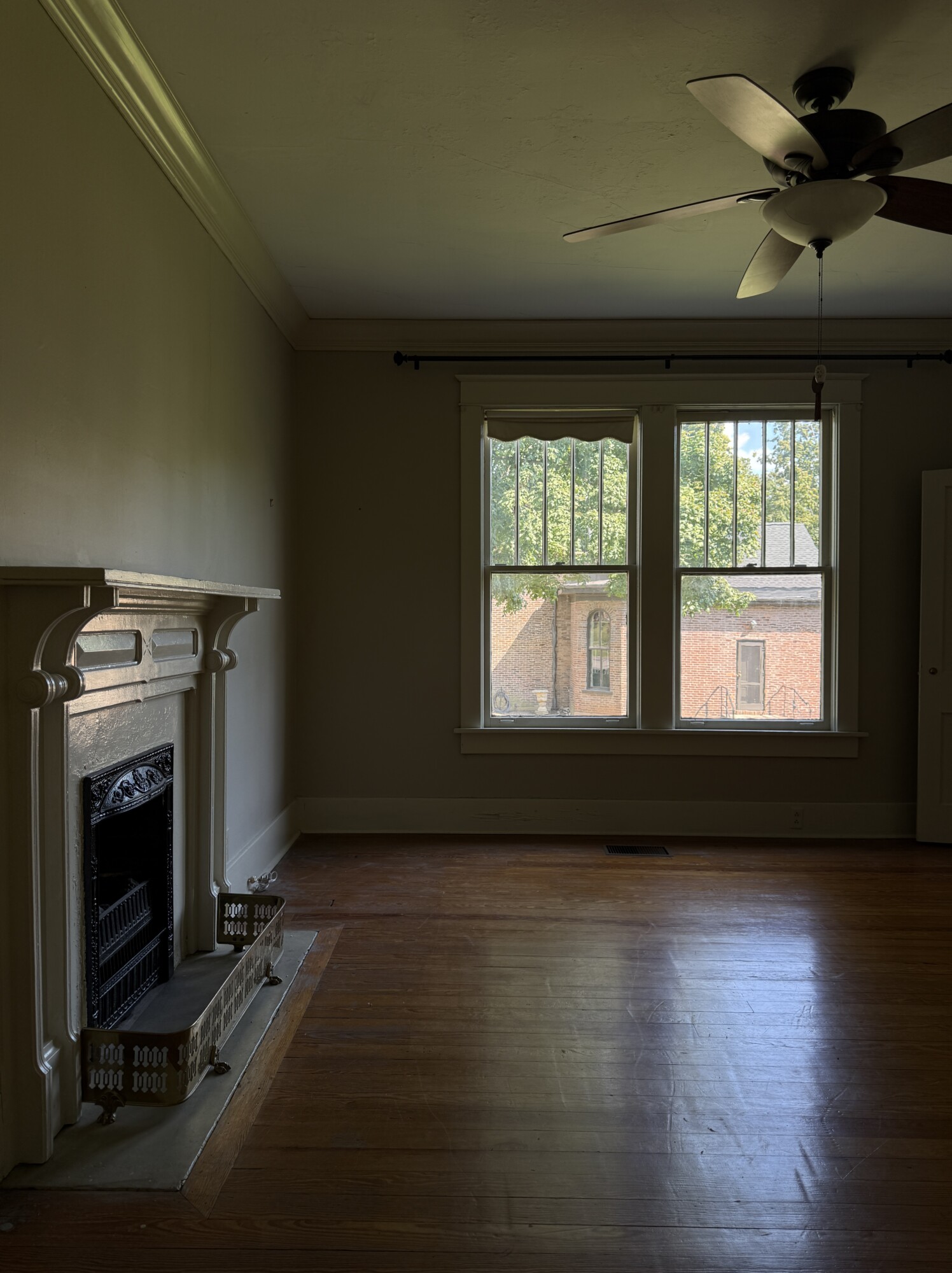 Front living room with ornate fireplace surround, large windows, ceiling fan, and worn hardwood floors — pre-renovation.