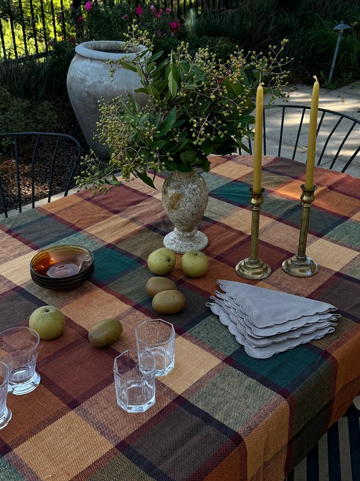 Outdoor dining table styled with a fall plaid tablecloth in warm tones, topped with a stone vase of greenery, brass candlesticks with taper candles, stacked linen napkins, pears, amber glass bowls, and clear drinking glasses, creating a cozy seasonal tablescape.