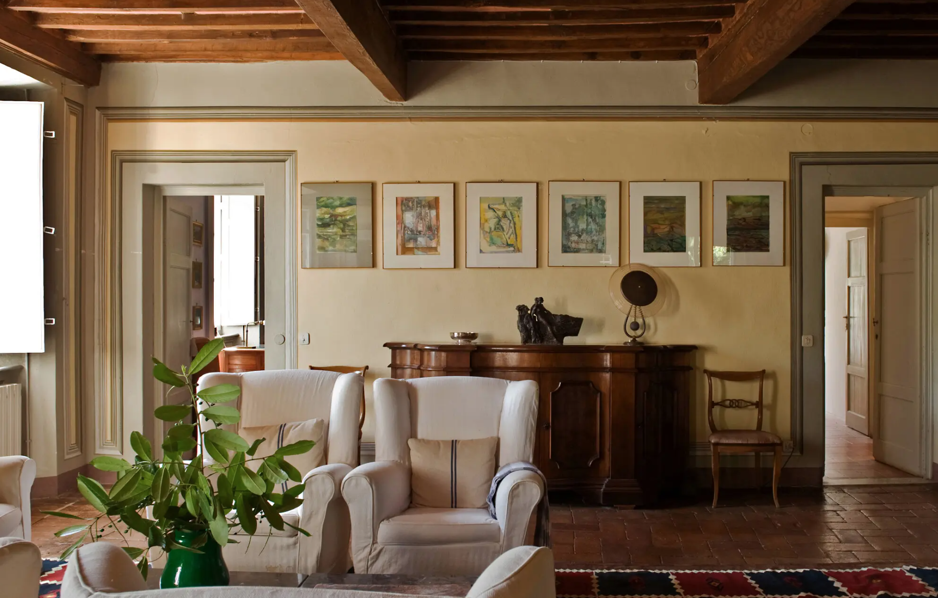 Sunlit Italian villa living room with exposed wood beams, a row of framed artworks above an antique sideboard, white slipcovered armchairs on terracotta tile floors, and a green vase of leafy branches on the coffee table beside open doorways.