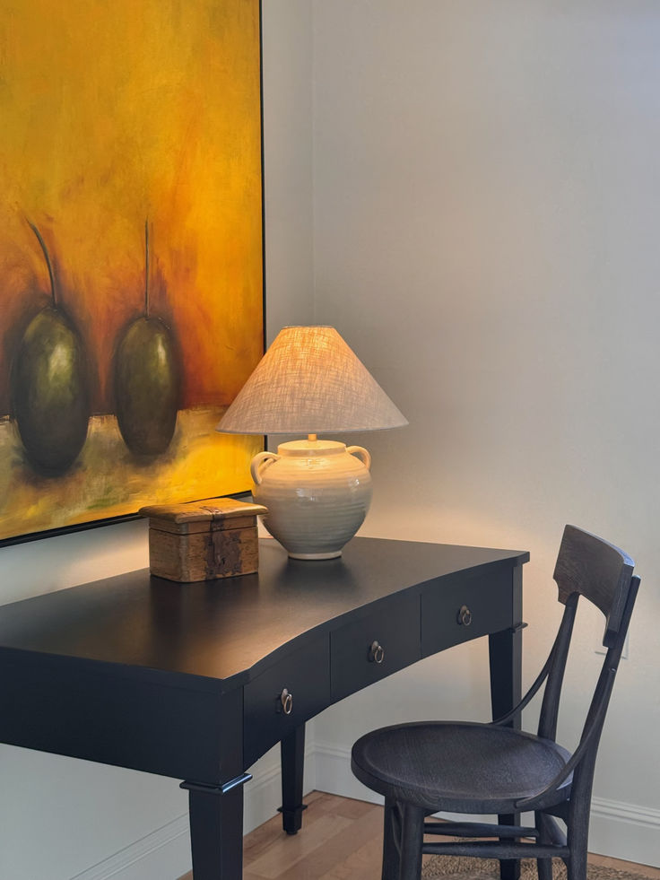 A cozy home office corner with a black writing desk, dark wood chair, and a ceramic jug lamp with a linen shade. On the desk sits a small wooden box, while a bold yellow and green abstract painting with two olives adds a dramatic backdrop.