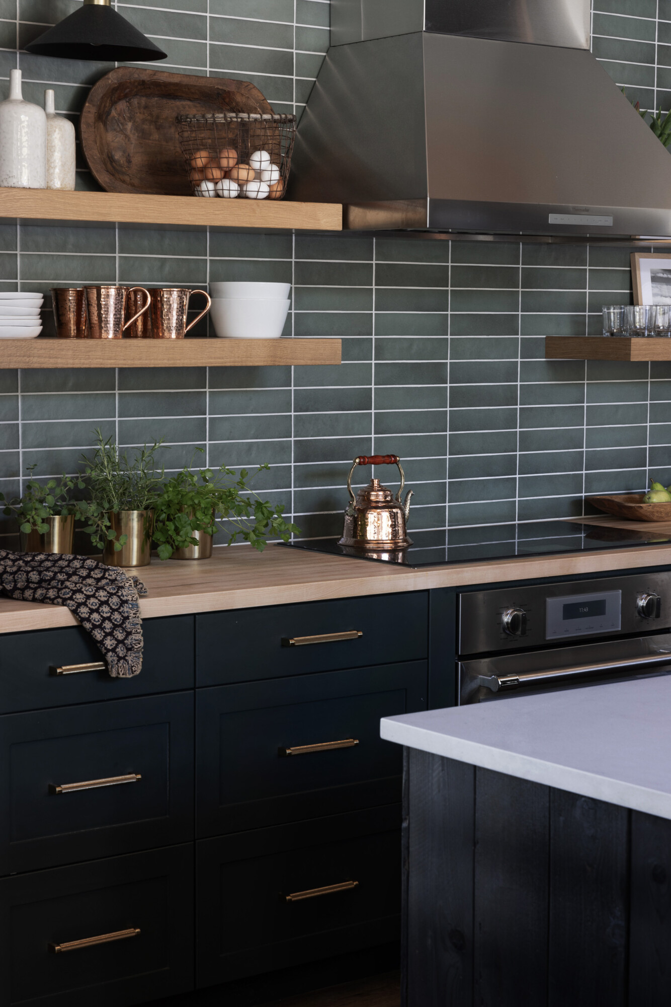 Modern kitchen with deep green cabinetry, brass hardware, and light wood countertops. Open wood shelves display copper mugs, white dishes, and a basket of eggs, while potted herbs in brass planters add freshness. A stainless steel range hood and copper kettle bring a mix of warmth and function against the green tiled backsplash.