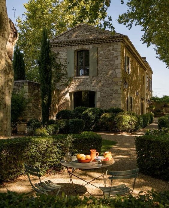 Sunlit stone country villa with green shutters and cypress trees, bordered by manicured hedges and a gravel path; in the foreground a round metal bistro table set with fruit, a carafe, and cups beneath leafy shade.
