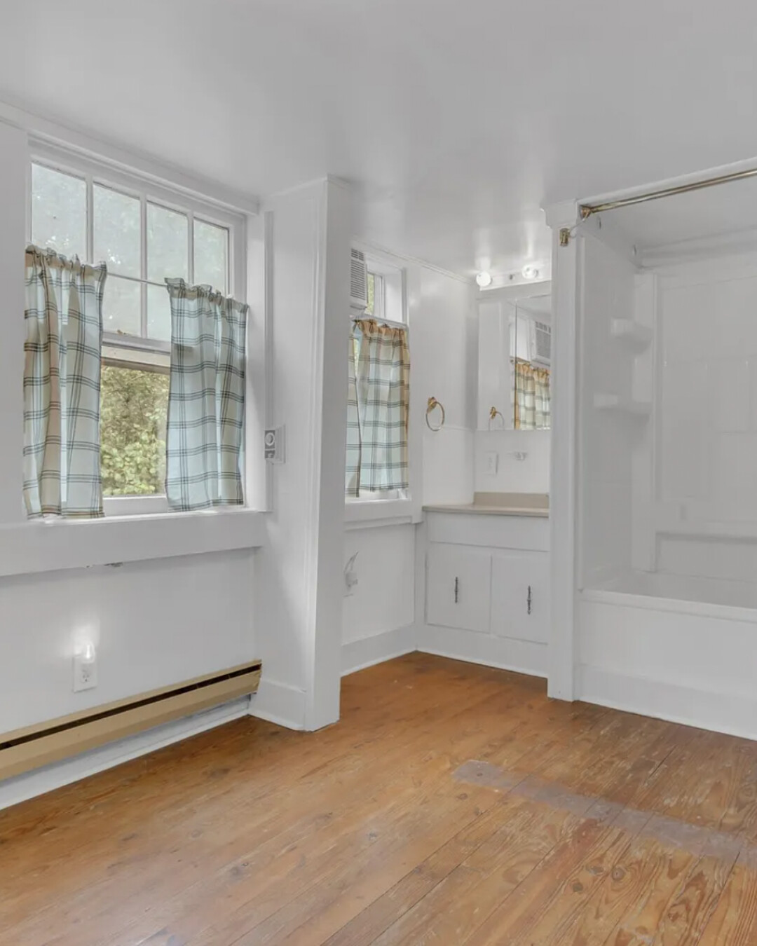 Bright, all-white bathroom with two tall windows, plaid café curtains, wood floors, and a built-in vanity; tub/shower combo at right.