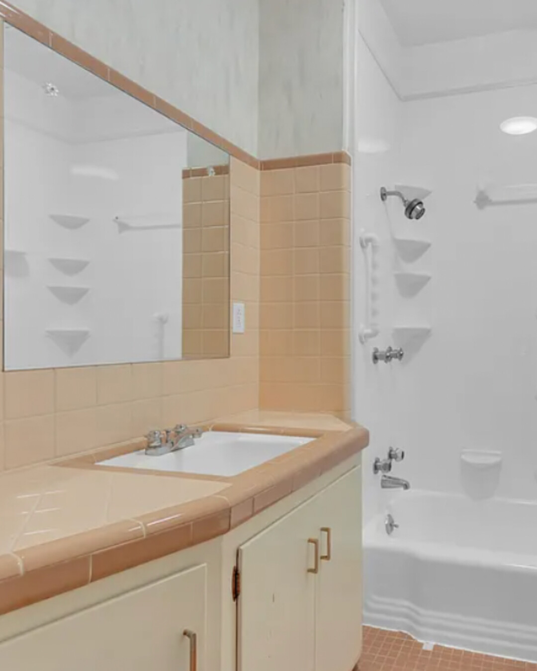 Vintage peach-tile bathroom vanity with integrated counter and backsplash, large wall mirror, and adjacent white tub/shower with corner shelves.