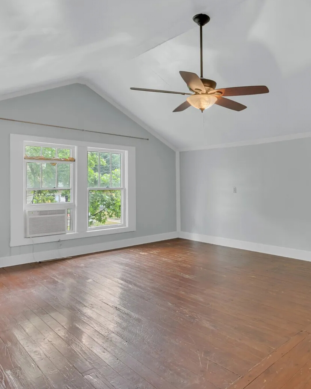 Spacious bedroom with vaulted white ceiling, gray walls, large double window with AC unit, wood plank floors, and a ceiling fan with light.