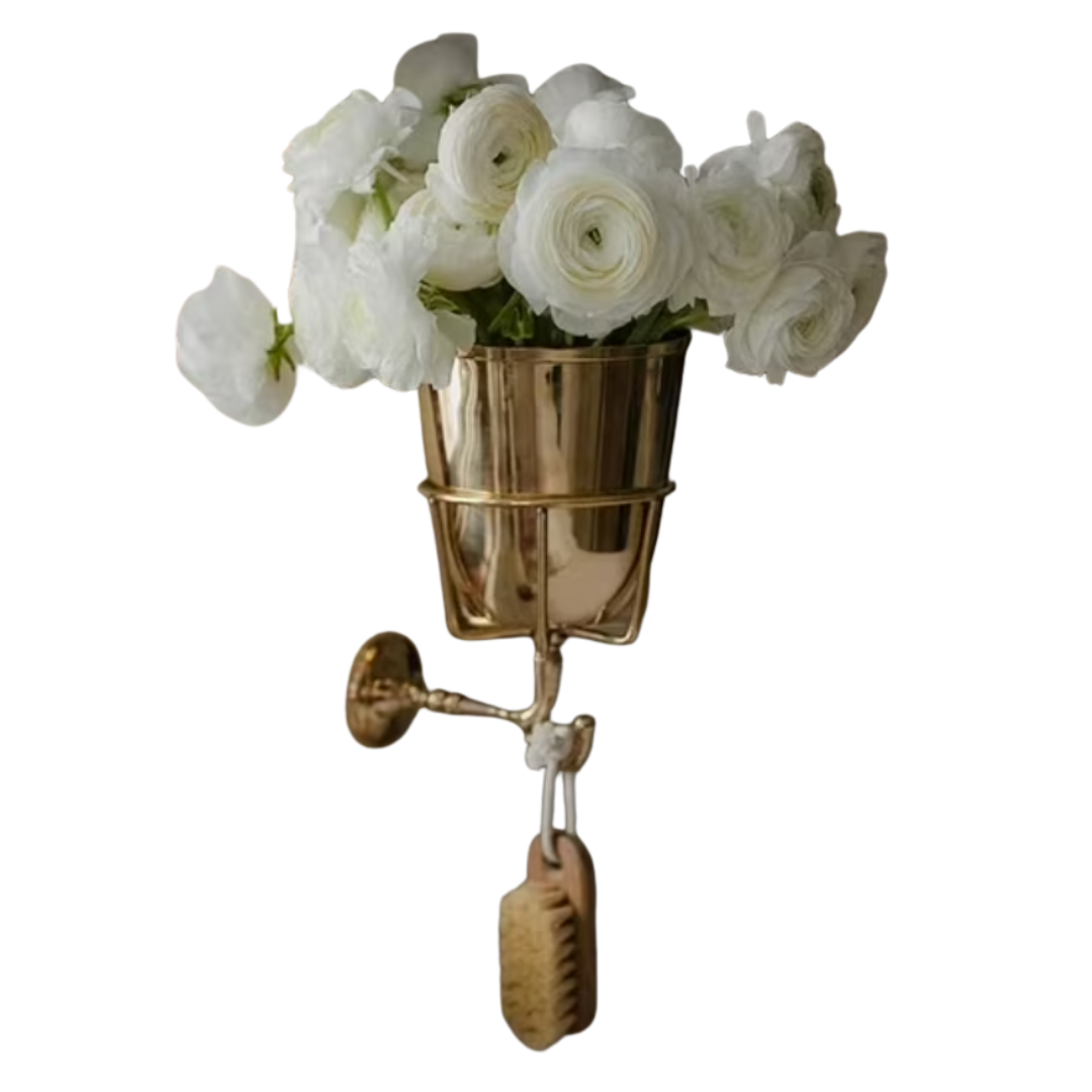 Wall-mounted polished brass cup/vase holding white ranunculus, hook for hanging brush.