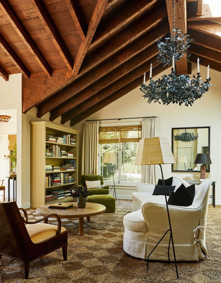 Warm living room with vaulted wood-beam ceiling, leafy black chandelier, built-in bookcase, large window with woven shades and drapery, white skirted sofas with black pillows, green velvet lounge chair, wood coffee table, patterned sisal-style carpet, and a slender iron floor lamp with a pleated shade
