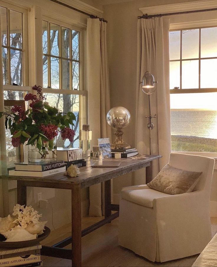 Warm sunlit study nook with a rustic wood desk by large paned windows overlooking water at sunset. Cream drapes, a white slipcovered chair, stacked books, a vase of flowers, and a glass globe lamp create a cozy, collected scene.