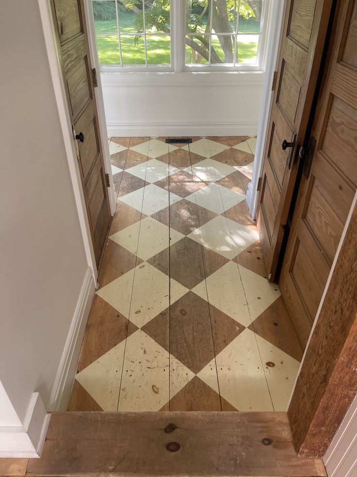 Sunlit hallway with a worn harlequin (diamond) painted pattern on wide-plank wood floors. Natural wood doors line both sides, leading to large windows that open to a green yard.