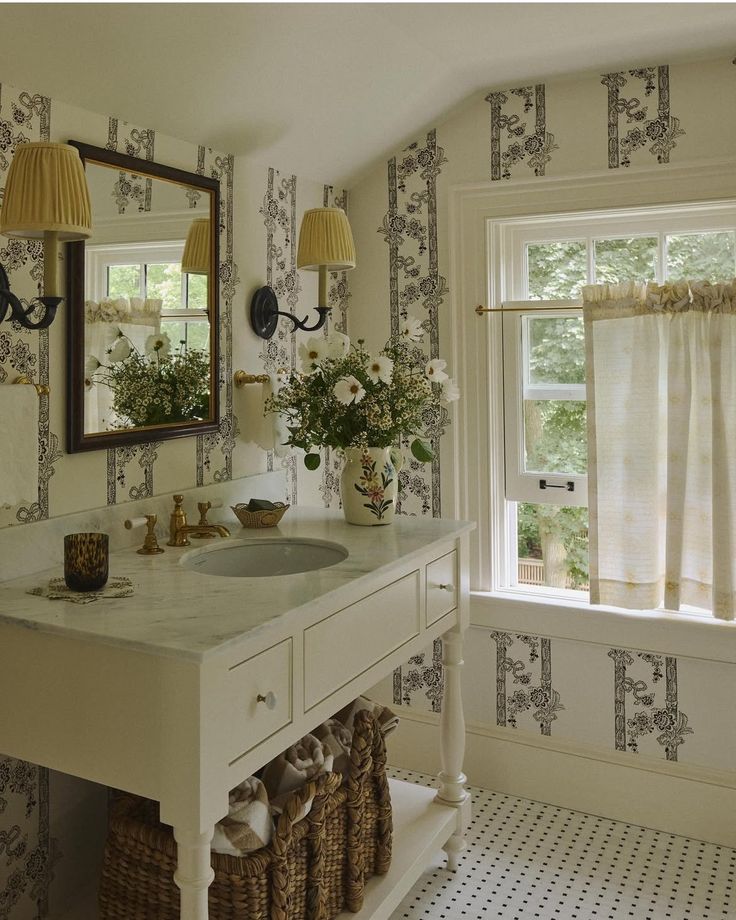 Cottage-style bathroom with patterned cream-and-charcoal wallpaper, a white marble-top vanity on turned legs, and brass faucet and hardware. Pleated yellow sconces flank a wood-framed mirror; a vase of white flowers, woven baskets on the lower shelf, café curtains, and tiny mosaic floor tiles complete the vintage look.