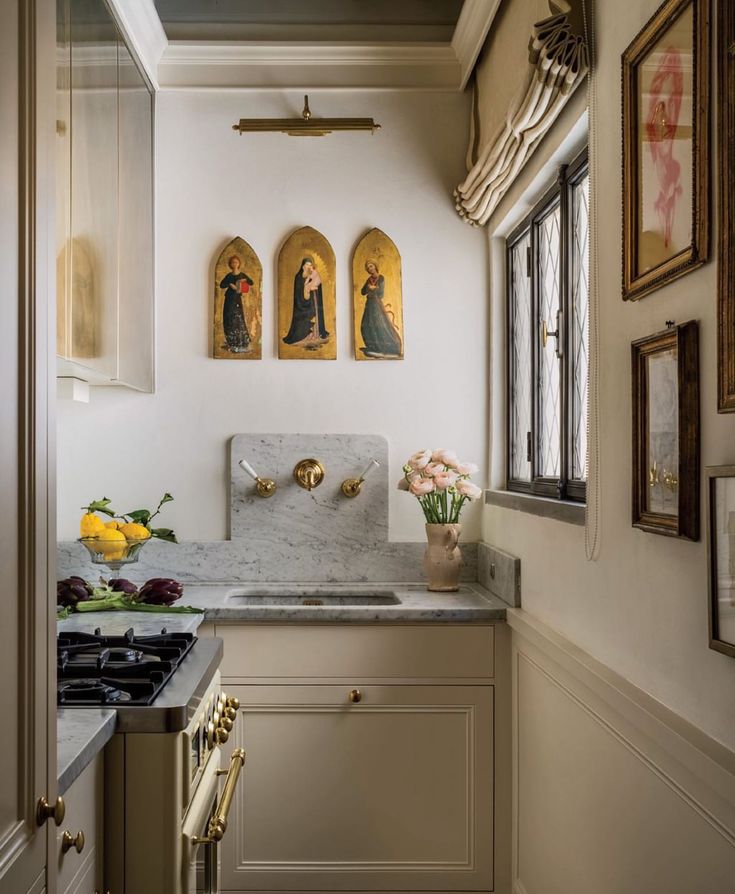 Small, elegant galley kitchen with creamy inset cabinets and marble counters; a marble slab backsplash holds a brass wall-mounted faucet. Above the sink, a trio of gilded medieval-style panels under a picture light, a pleated Roman shade over a leaded window, framed art along the wall, and styling details—lemons in a glass compote, pink flowers in a pottery vase, and a brass-trim range—create a collected, old-world feel.