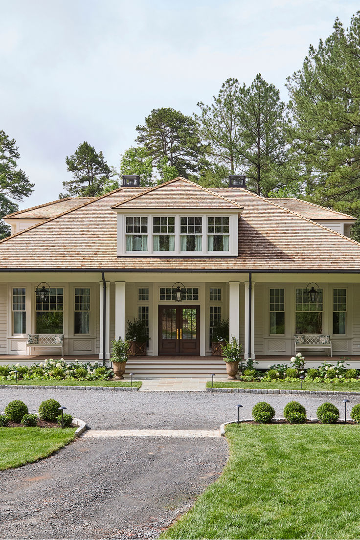 A symmetrical, light-taupe house with a cedar shingle hip roof and centered dormer. A deep front porch with columns and black lanterns overlooks flower beds, boxwood spheres, and a curved gravel drive bordered by lush lawn and tall pine trees.