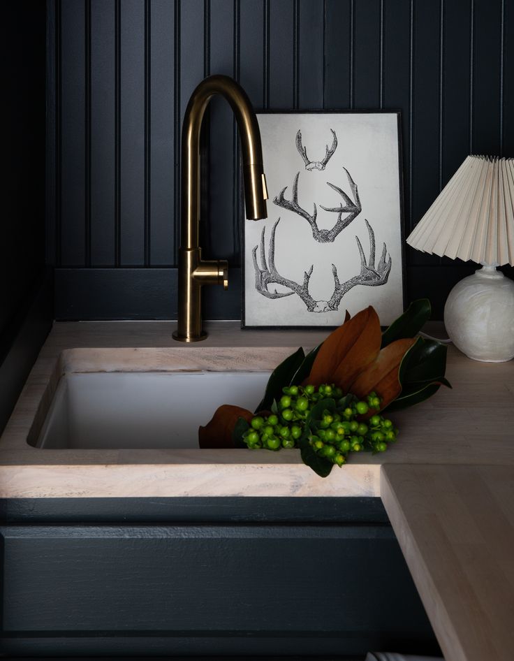 Moody laundry or prep sink vignette with a brass gooseneck faucet over a white inset sink in light butcher-block countertops, set against dark beadboard walls; framed antler art and a small pleated-shade lamp sit on the counter, with magnolia branches and green berries resting in the basin.