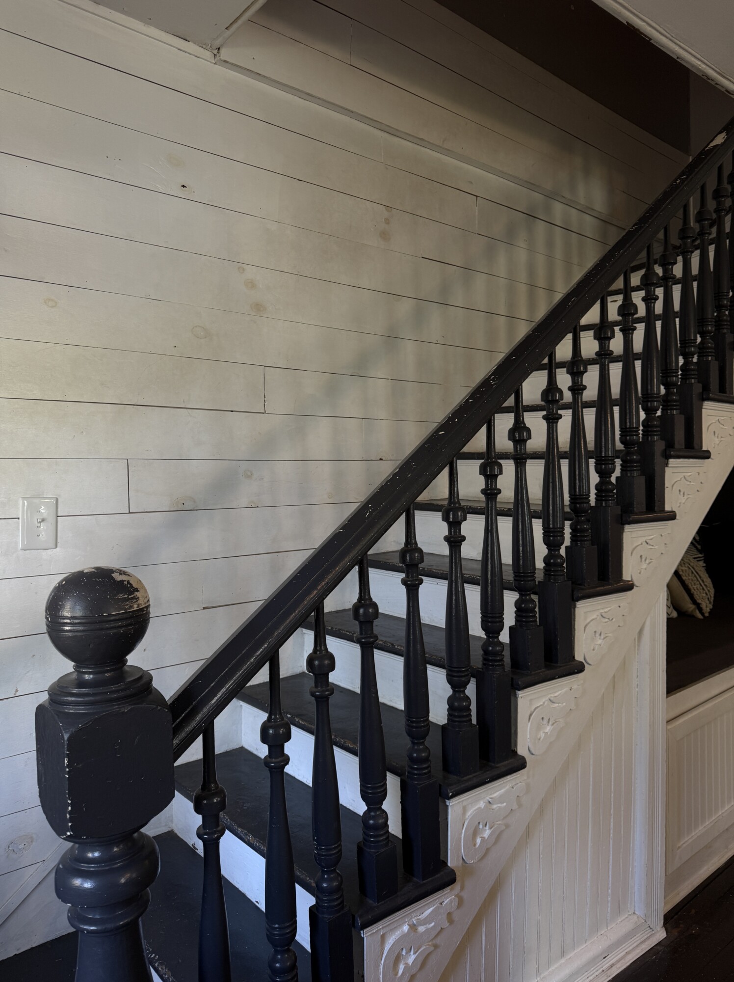 Interior staircase with glossy black balusters and newel post against white shiplap walls; worn treads and decorative stair brackets.