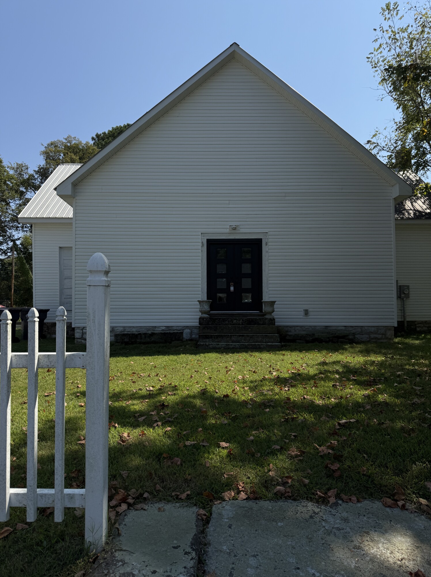 Front facade of a small white church building with gabled roof and black double doors, picket fence in the foreground.