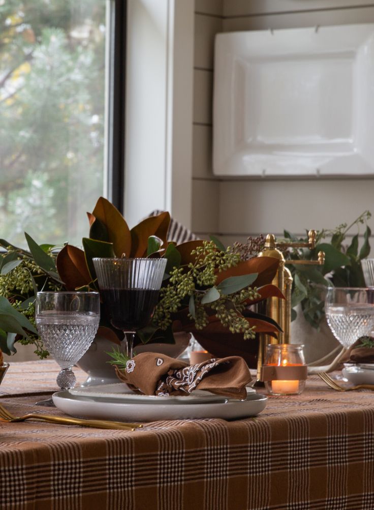 Thanksgiving tablescape with a brown plaid tablecloth, layered white plates, crystal goblets and a black wine glass, brass flatware, and a brown bandana napkin topped with a rosemary sprig; magnolia and eucalyptus centerpiece with votive candles by a window and shiplap wall with a hanging white platter.