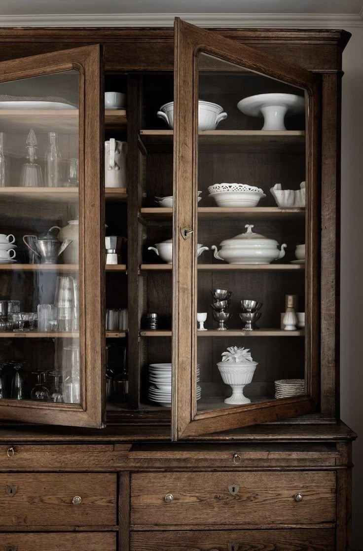 Antique oak glass-front hutch with one door ajar, showcasing neatly stacked white ironstone, silver goblets, and clear glassware on wood shelves. The lower cabinet features wide drawers with brass key escutcheons, all lit in warm, moody light for a collected, heirloom feel.