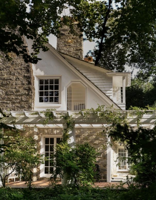 Sunlit stone-and-stucco cottage with a vine-draped pergola, white French doors and mullioned windows, steep gable and chimney, framed by lush garden greenery.