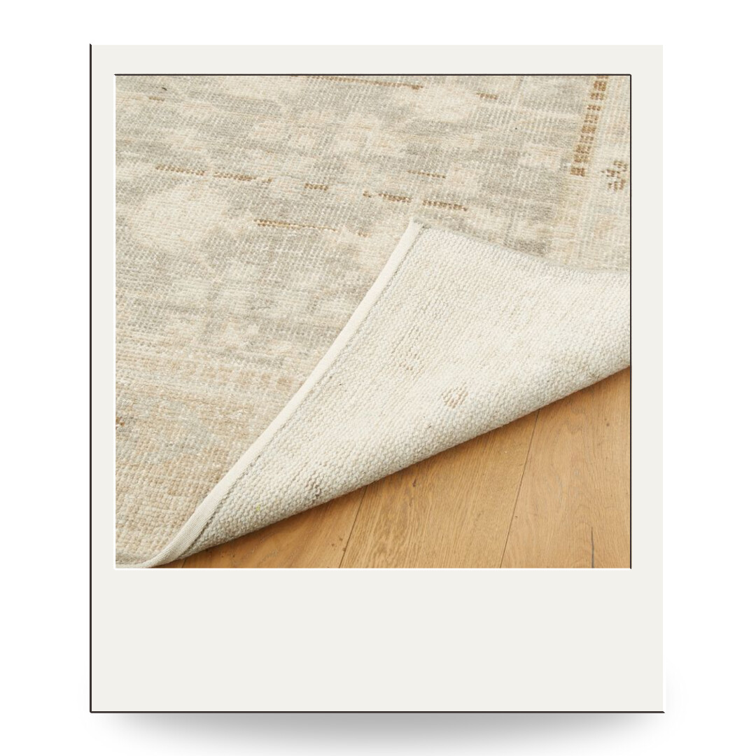 Neutral woven rug with a subtle faded pattern and texture detail, shown folded to reveal its backing.