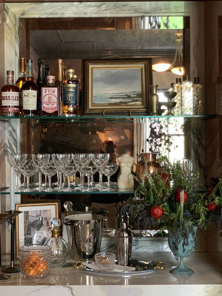 Elegant holiday bar setup with vintage glassware, liquor bottles, framed artwork, and a lush winter floral arrangement displayed on mirrored shelving.