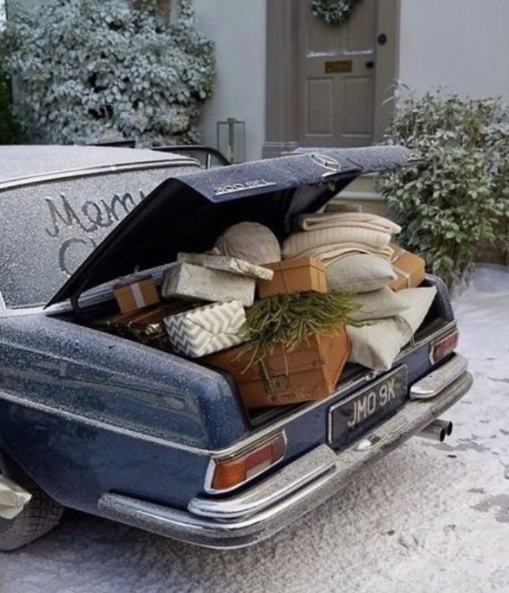 Trunk of a vintage blue car overflowing with wrapped Christmas presents, greenery, and cozy blankets, parked in front of a snow-dusted home with a wreath on the door. The scene captures the warmth and excitement of holiday travel and gift-giving on a snowy morning.