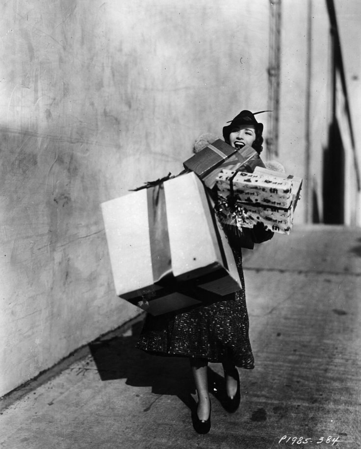 Black-and-white vintage photograph of a woman joyfully carrying a towering stack of wrapped Christmas presents down a city sidewalk. She’s dressed in a mid-century coat, hat, and heels, perfectly capturing the festive chaos and charm of holiday shopping in a bygone era.