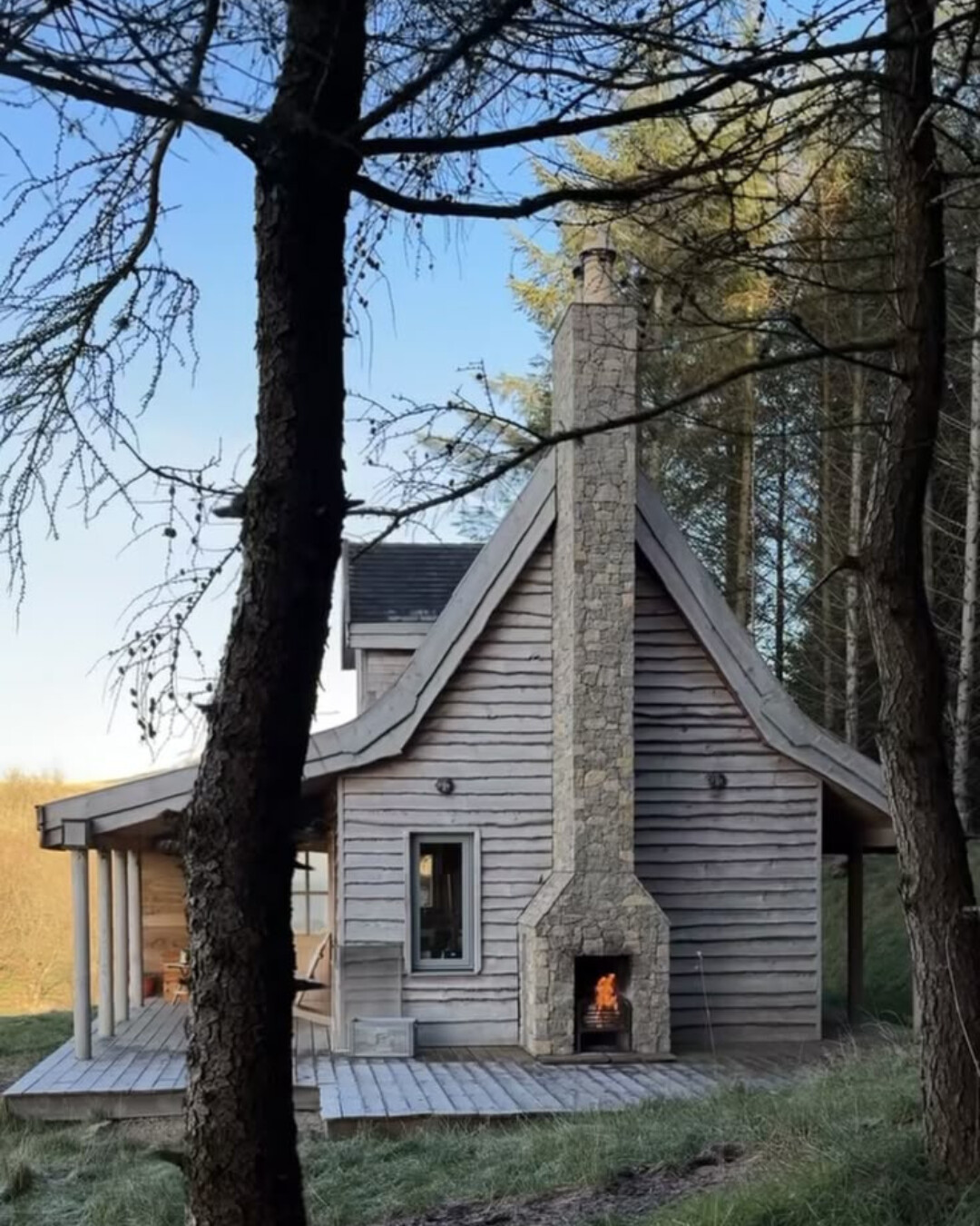 Cozy wood cabin with a tall stone chimney and outdoor fireplace, surrounded by trees and soft natural light, evoking a quiet winter retreat.