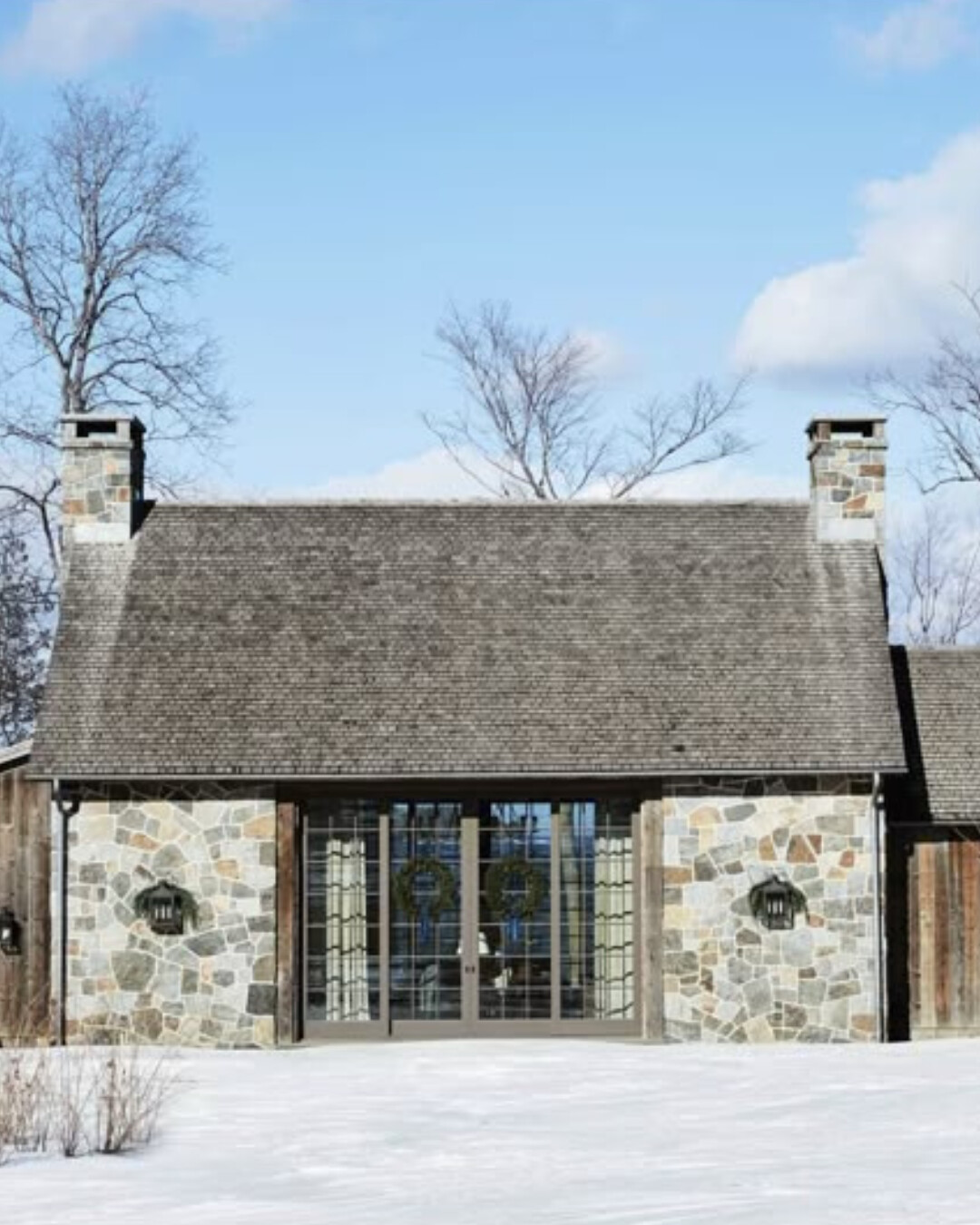 Stone cabin with wood accents and double glass doors framed by wreaths, set against a snowy landscape under a bright blue winter sky.