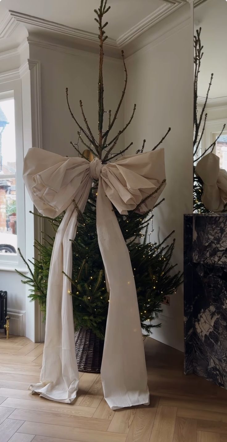 Minimal Christmas tree decorated with twinkle lights and an oversized ivory fabric bow, styled in a woven basket in a bright, modern room.