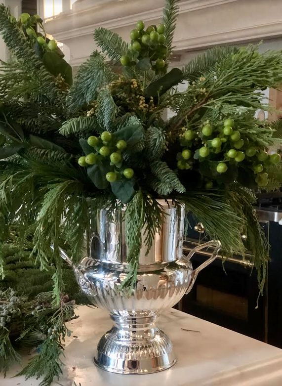 Large silver urn filled with fresh evergreen branches and green winter berries, styled as an elegant holiday centerpiece.