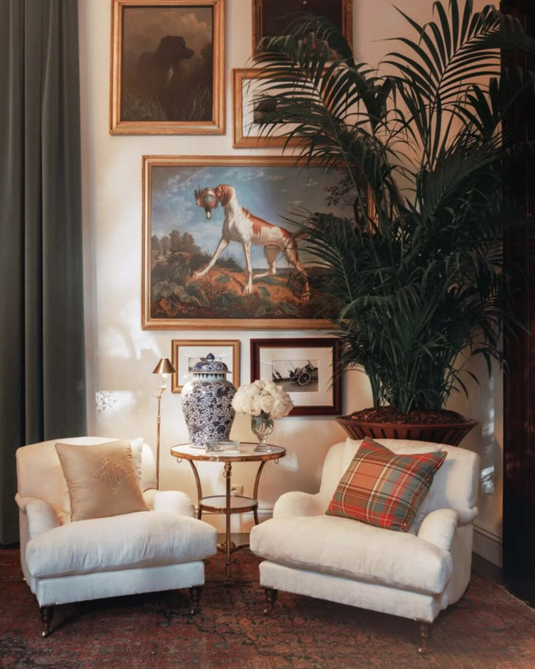 Two white upholstered lounge chairs sit in a traditional living room with a gallery wall of framed dog paintings. One chair has a tan monogrammed pillow, and the other has a classic red-and-green plaid pillow. A round brass side table is styled with a blue-and-white ginger jar and white flowers. A tall potted palm fills the corner behind the chairs.