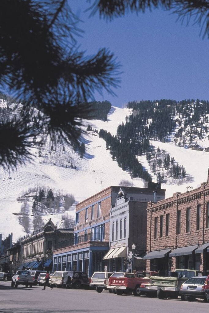 Downtown Aspen street view with snowy ski slopes rising behind historic buildings under a bright blue sky.