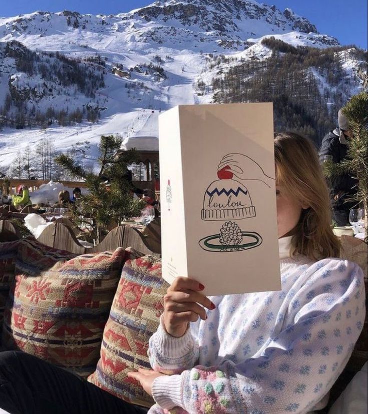 Woman relaxing outdoors in Aspen, holding up a menu with snowy mountains in the background and patterned pillows around her.
