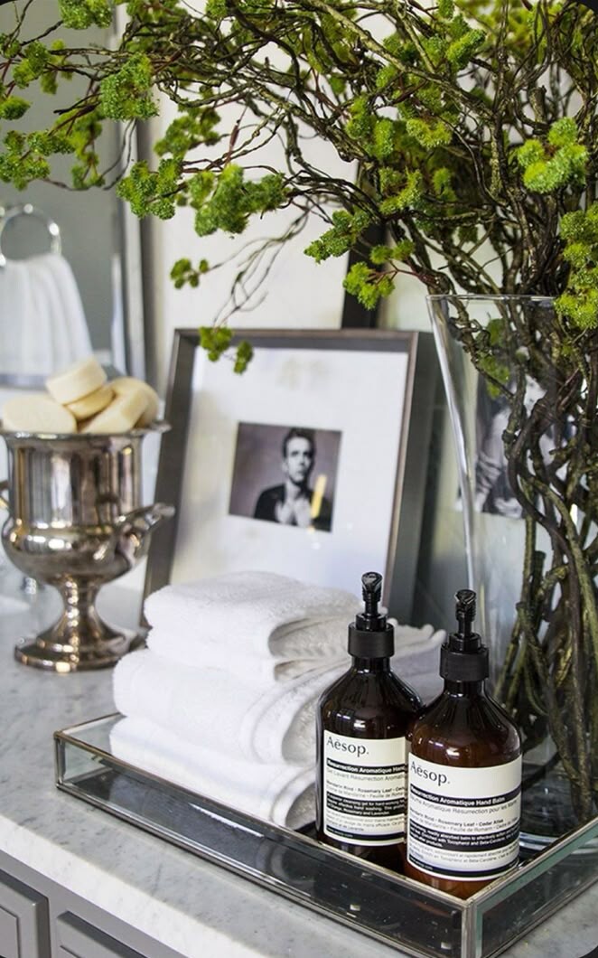 A bathroom or vanity countertop styled with stacked white towels on a rectangular glass tray beside two amber Aesop bottles. Behind them sits a framed black-and-white portrait leaning against the wall. To the left, a silver pedestal bowl holds several round soaps. On the right, tall branches with bright green buds are arranged in a clear glass vase, adding height and texture to the scene.