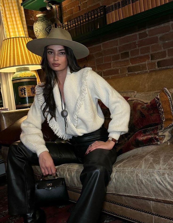 A woman in a white fringe sweater with black leather pants and a flat-brimmed cowboy hat. Sporting a polished Western look.