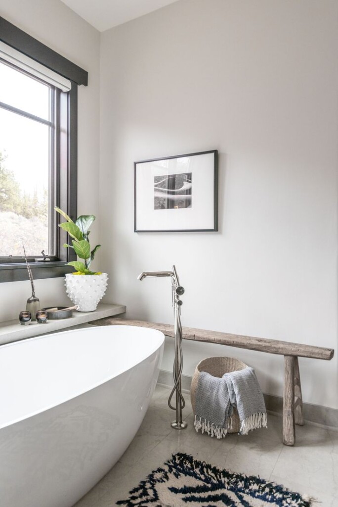 A modern neutral bathroom with a freestanding white soaking tub, a polished nickel floor-mounted tub filler, and a rustic wooden bench styled with a woven basket and textured throw. A white sculptural planter with greenery sits on the ledge beneath a black-framed window, and a simple black-and-white photo hangs above, creating a calm and minimalist spa-like atmosphere.