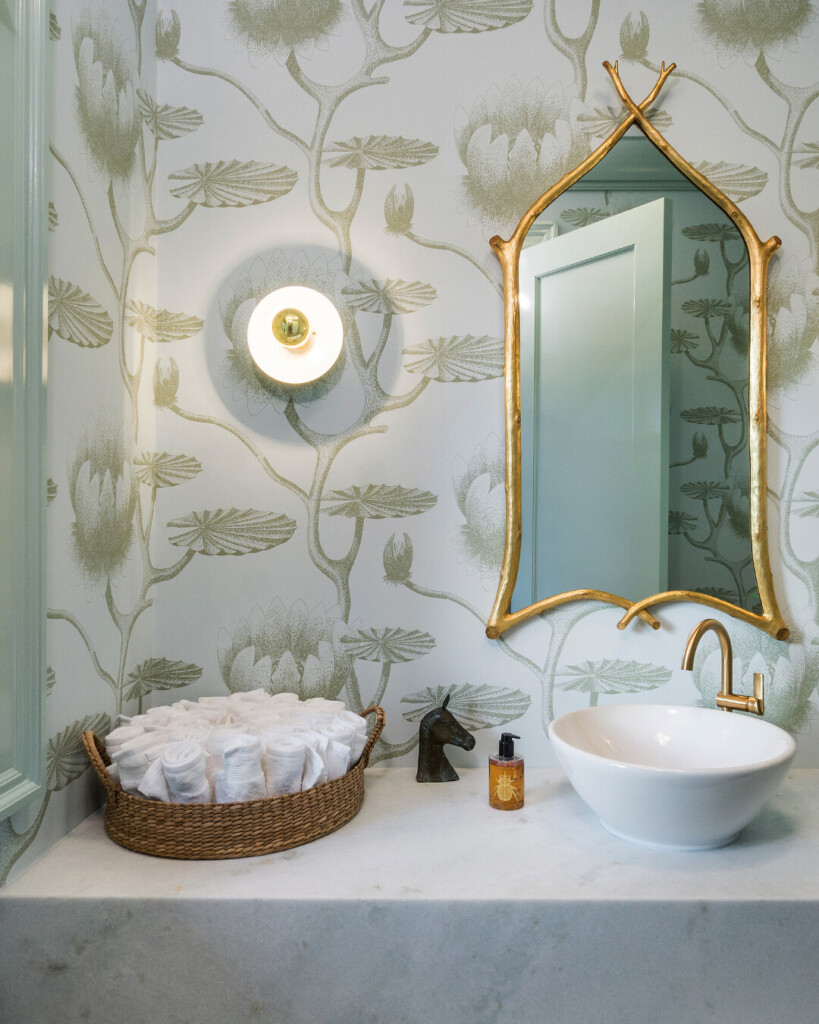 A bathroom vanity with a marble countertop, a round white vessel sink, and a gold faucet. A gold-framed mirror with an arched, branch-like shape hangs on the wall above the sink. A modern sconce with a brass center is mounted to the left. On the counter sits a woven basket filled with rolled white washcloths, a small bronze horse-head figurine, and an amber-colored hand soap bottle. The walls are covered in a botanical wallpaper with large lotus-style flowers in soft green tones.