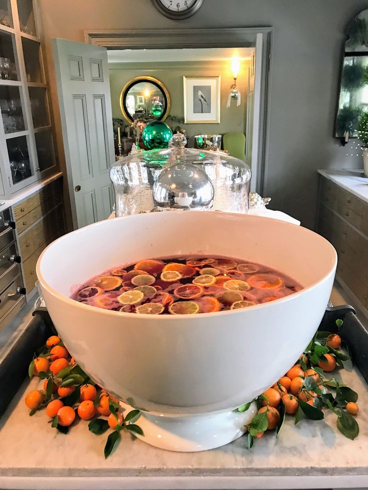A large white ceramic punch bowl filled with a red holiday punch topped with floating citrus slices—blood orange, lemon, and lime. The bowl sits on a marble tray surrounded by fresh orange branches with leaves. In the background is a warmly lit kitchen with gray cabinetry, glassware, and a doorway leading to another decorated room with green ornaments and a round mirror.