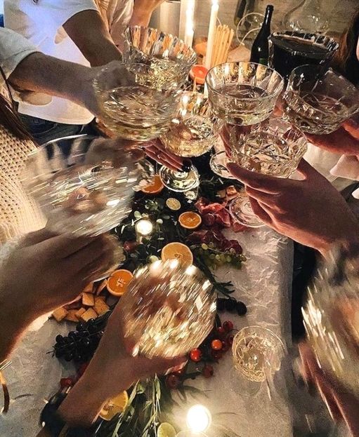 Several people raise crystal coupe glasses in a toast over a candlelit table. The table is set with a charcuterie-style spread of sliced citrus, grapes, cheeses, and greenery. Warm candlelight glows across the scene, creating a festive, celebratory atmosphere.