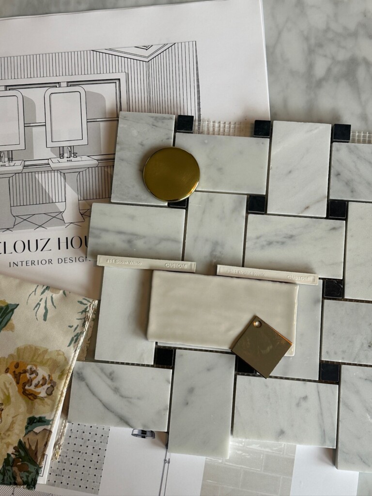 Close-up of marble tile samples laid out in a basketweave pattern, comparing two grout color options with brass hardware accents placed on top to show subtle differences in tone and contrast.