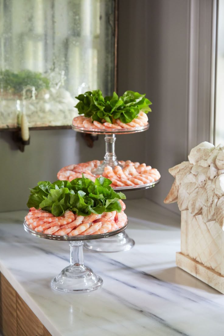 A tiered display of chilled shrimp rings arranged on clear glass pedestal stands, each topped with a bundle of fresh green lettuce leaves in the center. The stands sit on a marble countertop near an antique-style distressed mirror, creating an elegant, classic presentation.