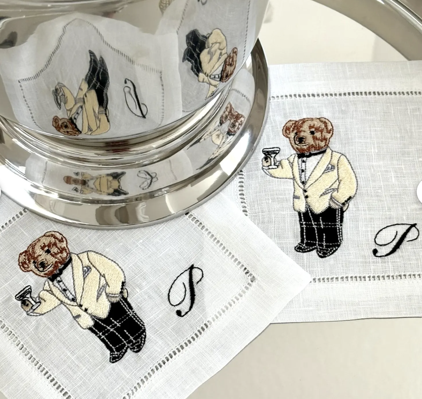 A close-up of two white linen cocktail napkins embroidered with a tuxedo-clad Polo Bear holding a martini glass, each napkin featuring a black stitched monogram “P.” The napkins sit beneath and beside a shiny silver ice bucket, which reflects the bear embroidery in its polished surface.