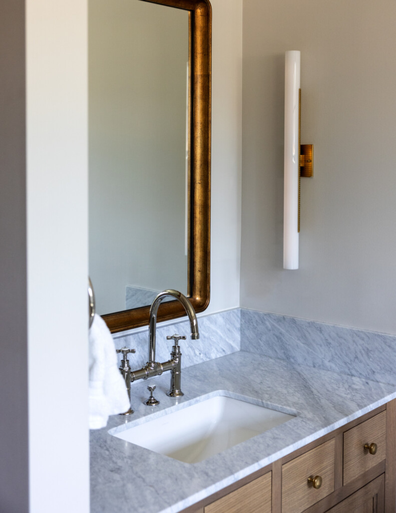 A bathroom vanity featuring a polished nickel bridge faucet mounted on a honed marble countertop. A large antiqued brass-framed mirror hangs above the sink, paired with a sleek vertical white sconce on the adjacent wall. Warm wood drawers with brass knobs add contrast against the cool marble, creating a refined and timeless look.