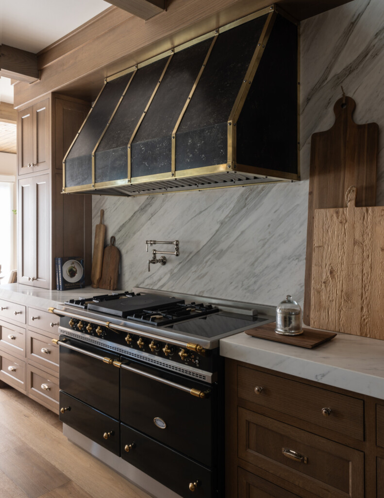 Close-up view of a luxury kitchen range area featuring a black and brass Ilve-style stove beneath a large custom range hood with black panels and brass trim. The backsplash is a slab of soft gray-veined marble, with a polished nickel pot filler mounted above the cooktop. Warm wood cabinetry with brass hardware surrounds the space, along with wood cutting boards and a small marble cloche displayed on the counter. The overall look feels elevated, timeless, and warmly traditional.