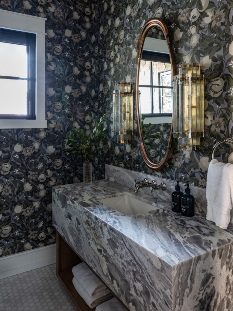 Moody powder bathroom with dark botanical wallpaper and a thick, waterfall-style marble vanity in gray and white tones. A polished nickel wall-mounted faucet sits above an undermount sink, with two vintage-inspired glass sconces flanking an oval bamboo-style mirror. Natural light enters through a black-trimmed window, and folded white towels are stored on the open wood shelf beneath the vanity.