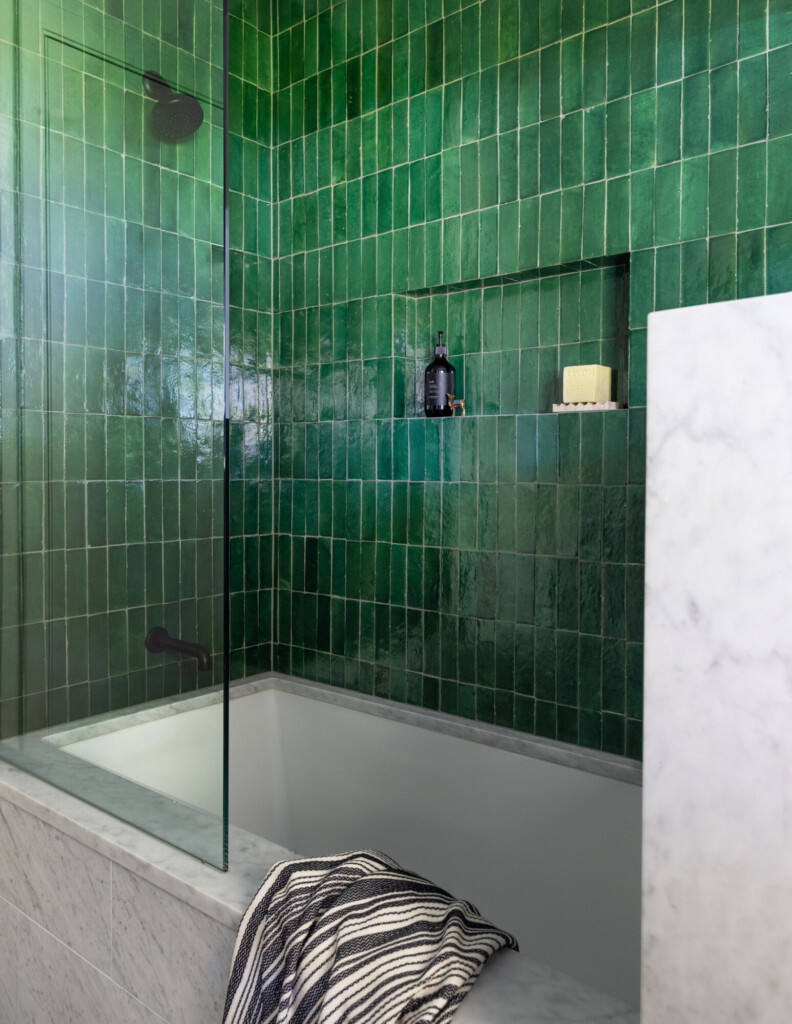 Bathroom with deep green zellige tile walls, a built-in shower niche, matte black fixtures, and a marble-surround bathtub with a striped towel draped over the edge.