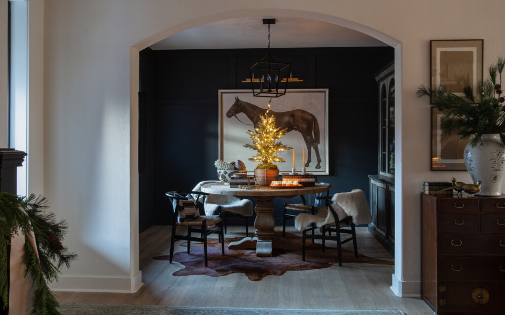 Moody dining room with dark walls, horse artwork, round wood table, and a mini lit Christmas tree centerpiece.